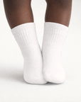 Baby with a dark skin tone wearing a pair of white baby crew socks on an off-white background.