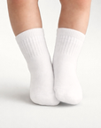 Baby with a light skin tone wearing a pair of white baby crew socks on an off-white background.
