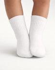 Baby with a medium skin tone wearing a pair of white baby crew socks on an off-white background.