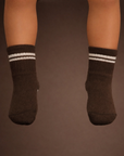 Baby with a medium skin tone wearing a pair of espresso brown baby crew socks with white heart and dot patterned silicone grips on an espresso brown background