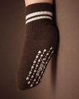 Baby with a light skin tone wearing an espresso brown baby crew sock with white heart and dot silicone grips on an espresso brown background