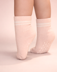 Baby with a light skin tone wearing a pair of blush baby crew socks with white heart and dot patterned silicone grips on a blush background