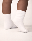 Baby with a dark skin tone wearing a pair of white baby crew socks with heart and dot patterned white silicone grips on an off-white background
