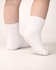 Baby with a light skin tone wearing a pair of white baby crew socks with heart and dot patterned silicone grips on an off-white background