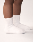 Baby with a dark skin tone wearing a pair of white baby crew socks with silicone grips on an off-white background