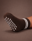 Baby with a medium skin tone wearing an espresso brown baby crew sock with white heart and dot silicone grips on an espresso brown background