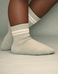 Baby with a dark skin tone wearing a pair of sage baby crew socks with silicone grips on a sage background