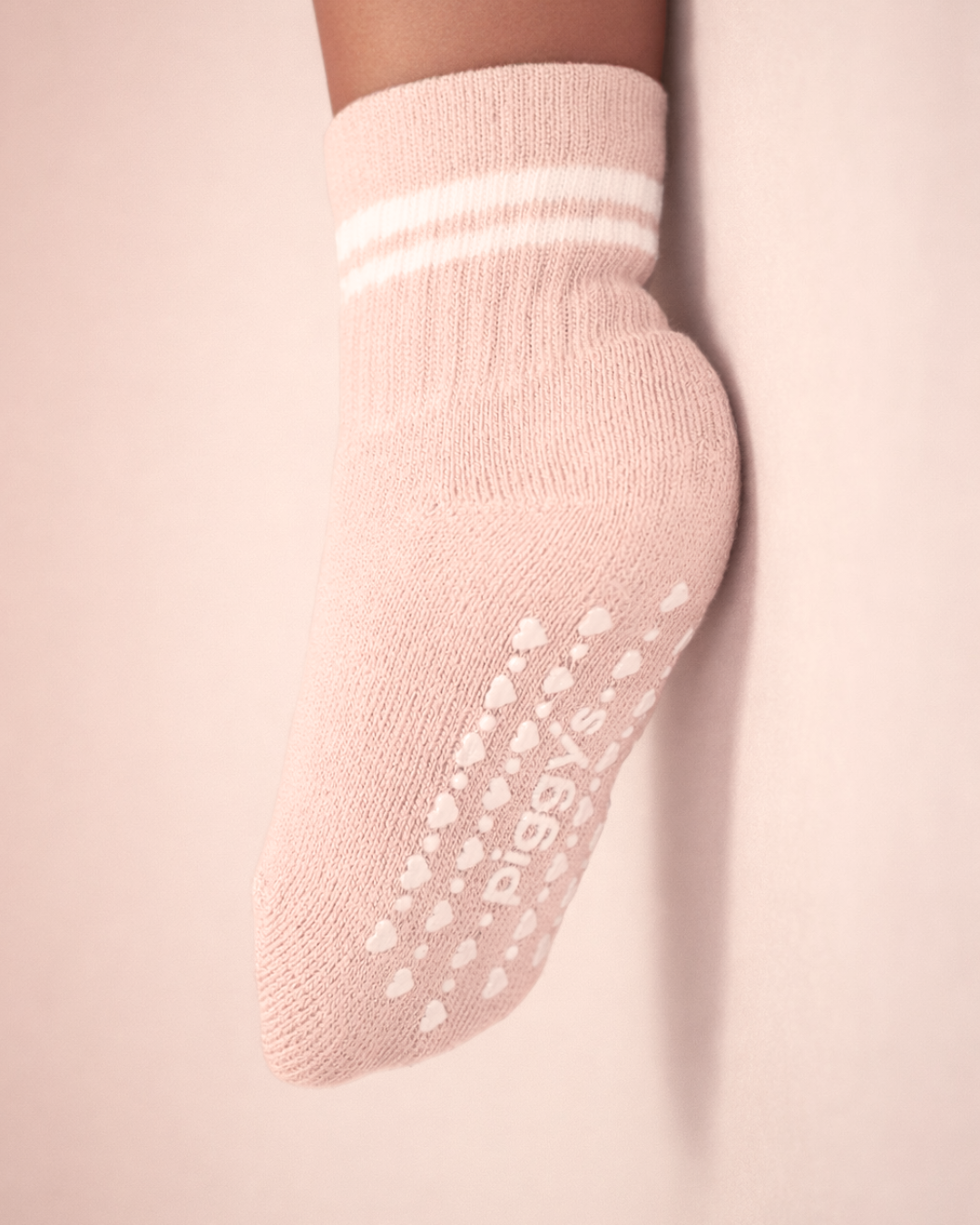Baby with a medium skin tone wearing a blush baby crew sock with white heart and dot silicone grips on a blush background