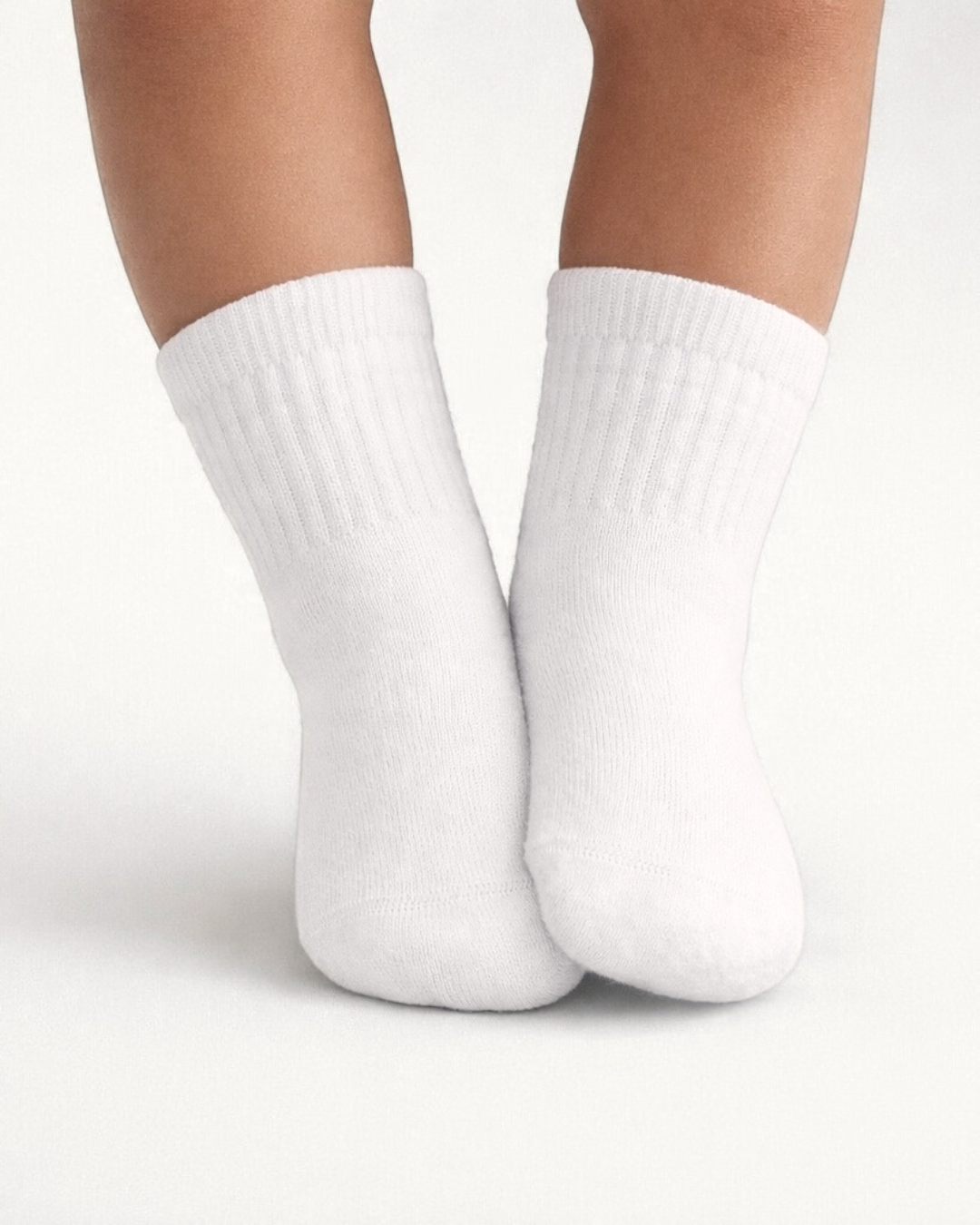 Baby with a medium skin tone wearing a pair of white baby crew socks on an off-white background.