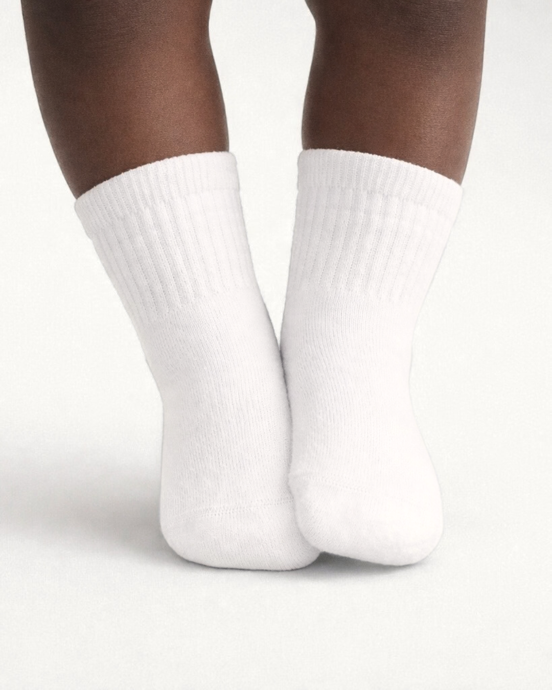 Baby with a dark skin tone wearing a pair of white baby crew socks on an off-white background.