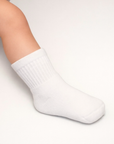 Baby with a light skin tone wearing a pair of white baby crew socks on an off-white background.