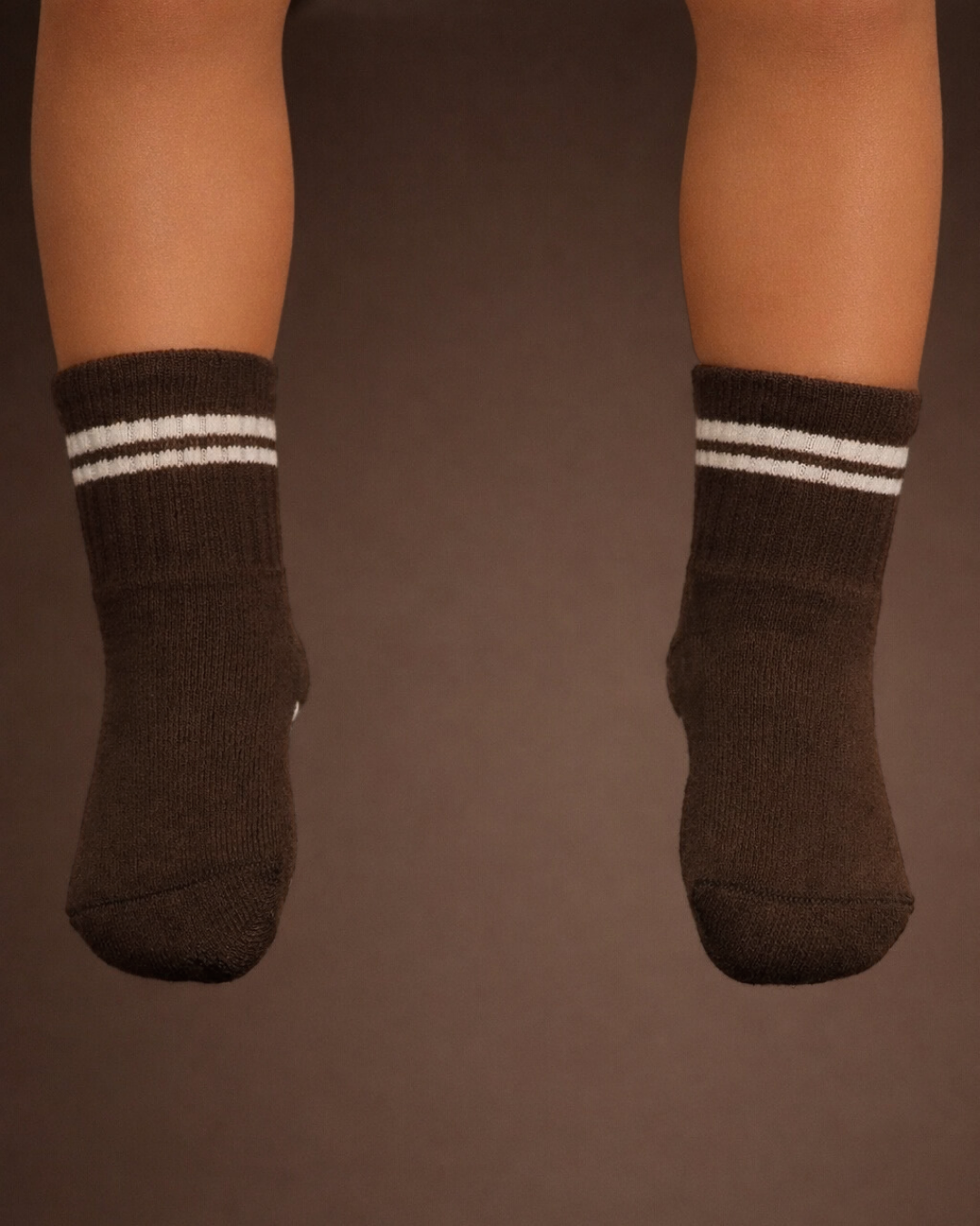 Baby with a medium skin tone wearing a pair of espresso brown baby crew socks with white heart and dot patterned silicone grips on an espresso brown background