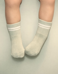 Baby with a light skin tone wearing a pair of sage baby crew socks on a sage background.