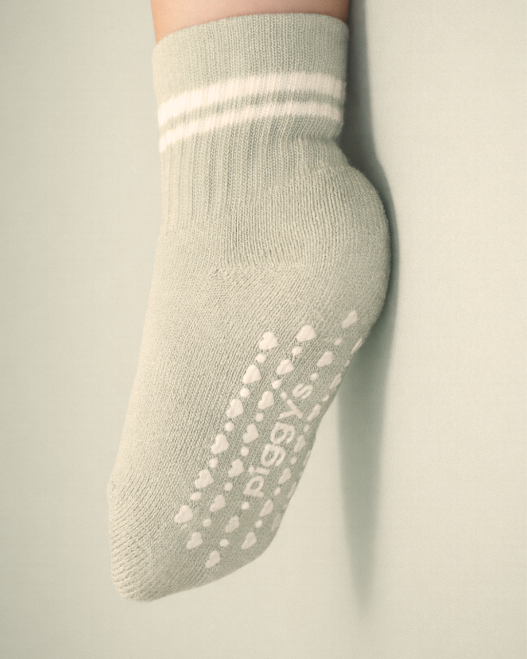 Baby with a light skin tone wearing a sage baby crew sock with white heart and dot silicone grips on a sage background