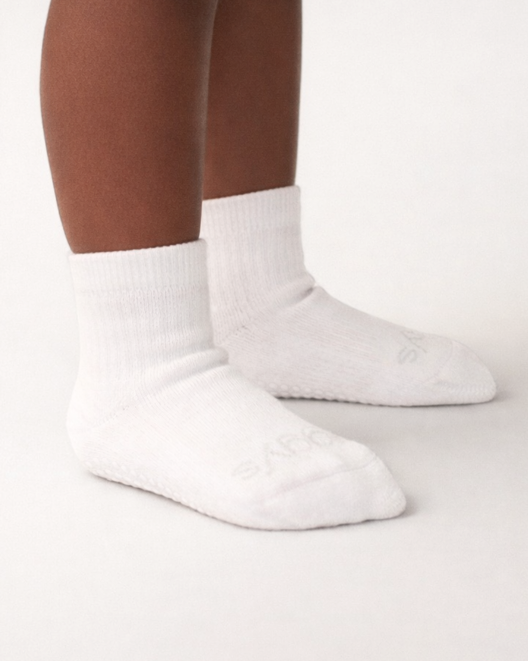 Toddler with a dark skin tone wearing a pair of white toddler crew socks on an off-white background.