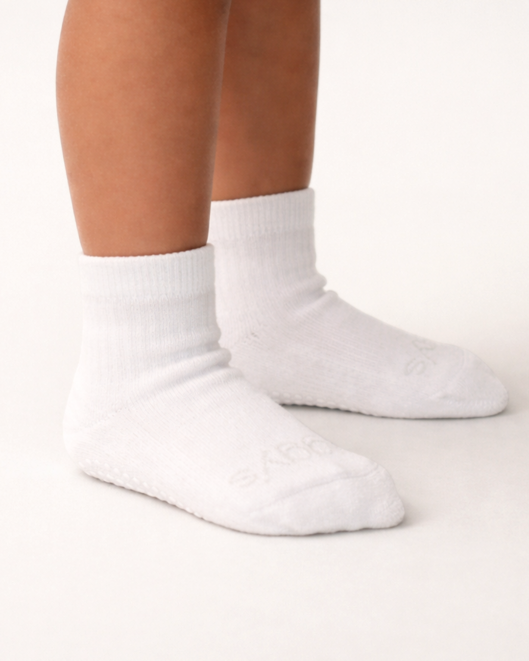 Toddler with a medium skin tone wearing a pair of white toddler crew socks on an off-white background.