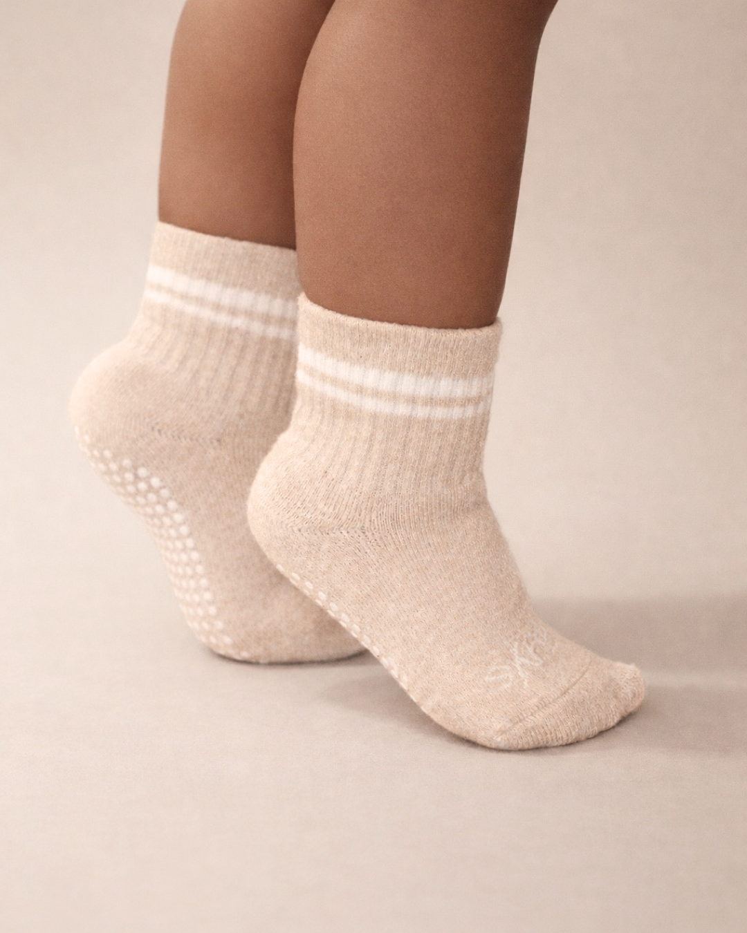 Toddler with a medium skin tone wearing a pair of heather beige toddler crew socks on a beige background.