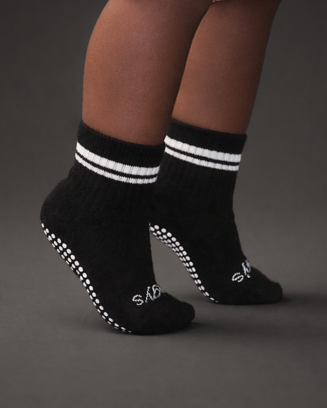 Toddler with a dark skin tone wearing a pair of black toddler crew socks on a dark grey background.