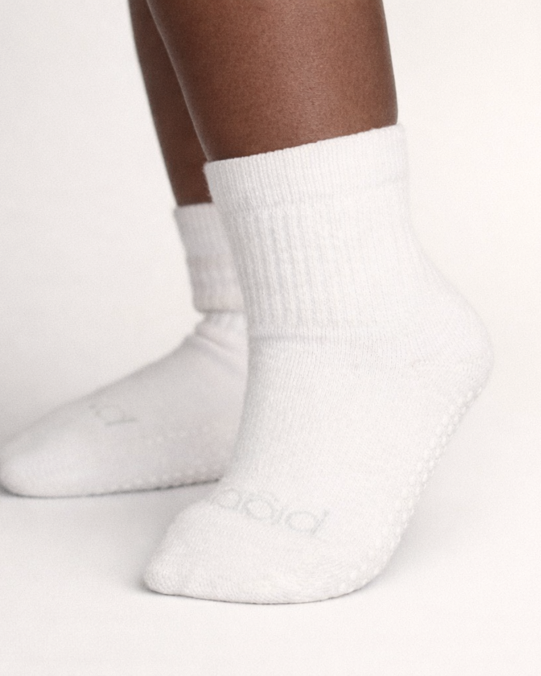 Toddler with a dark skin tone wearing a pair of white toddler crew socks on an off-white background.