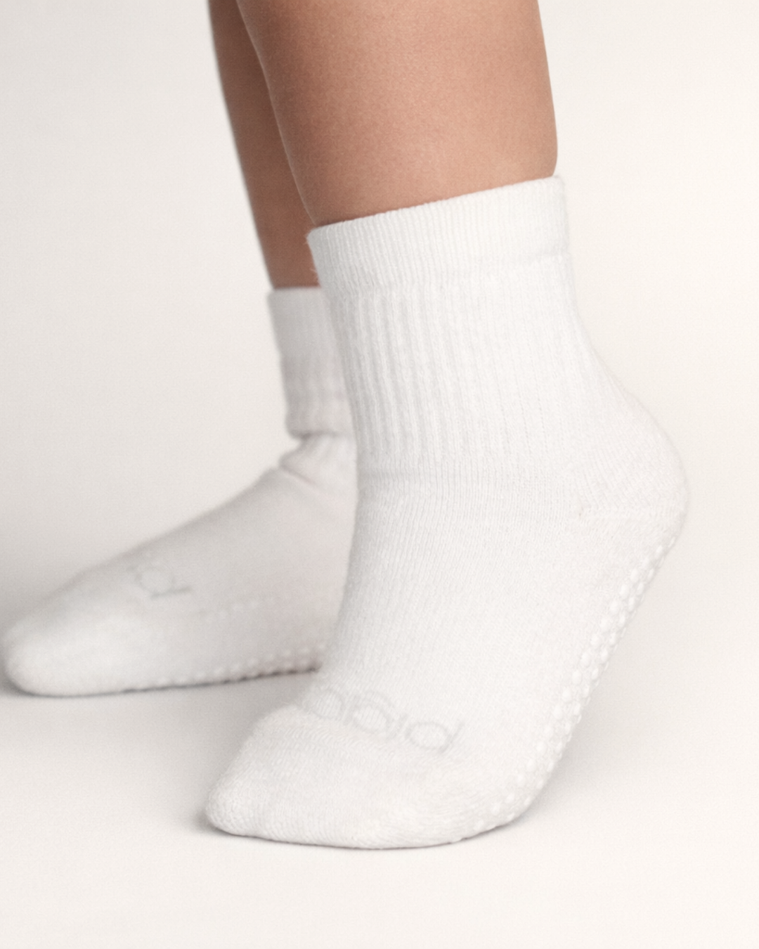 Toddler with a light skin tone wearing a pair of white toddler crew socks on an off-white background.