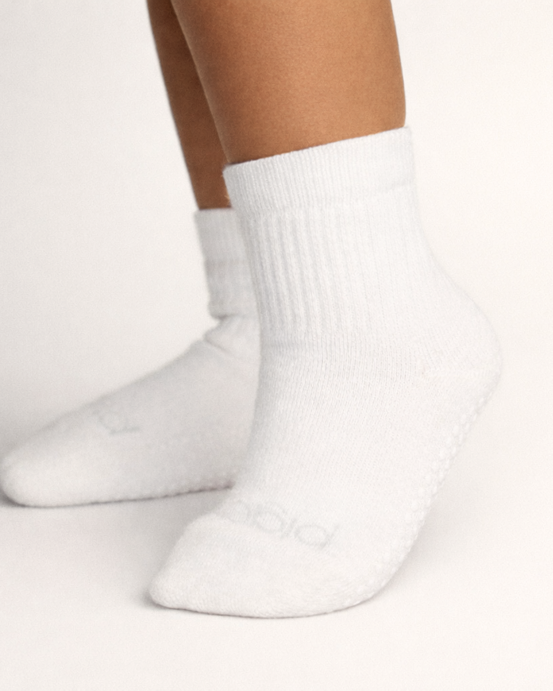 Toddler with a medium skin tone wearing a pair of white toddler crew socks on an off-white background.
