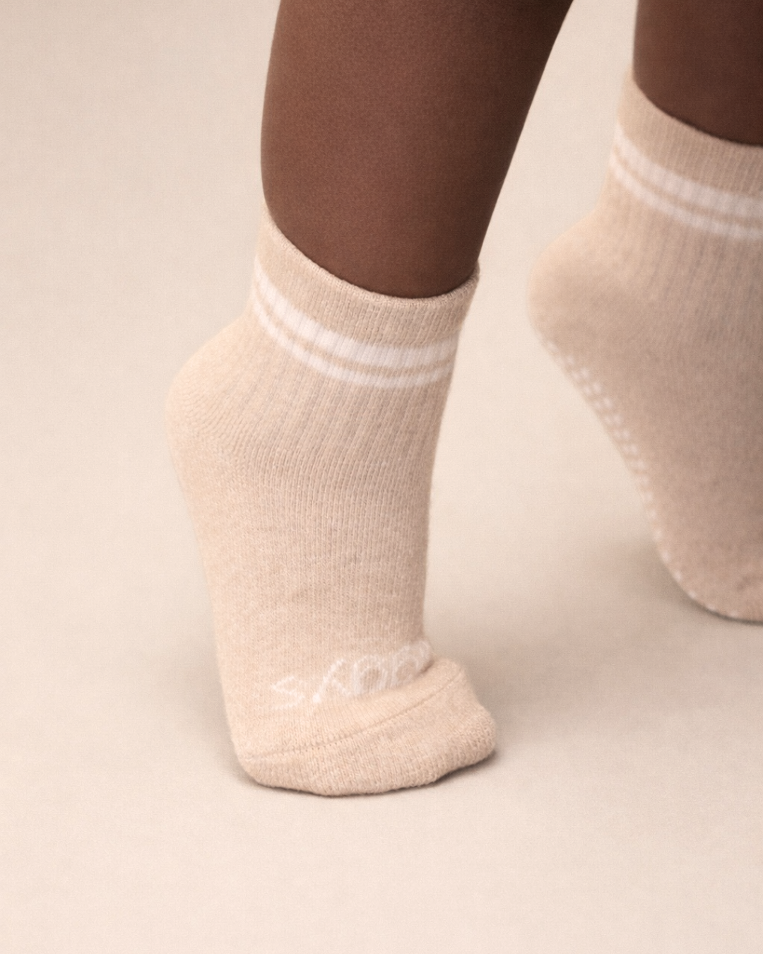 Toddler with a dark skin tone wearing a pair of heather beige toddler crew socks on a beige background.