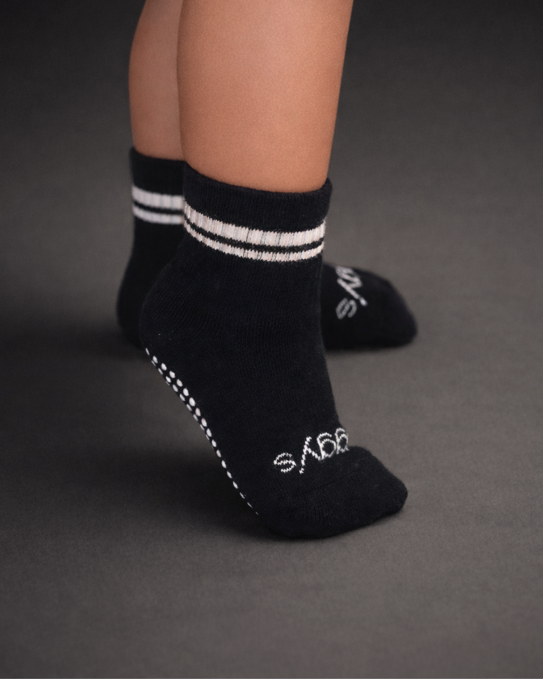 Toddler with a medium skin tone wearing a pair of black toddler crew socks on a dark grey background.