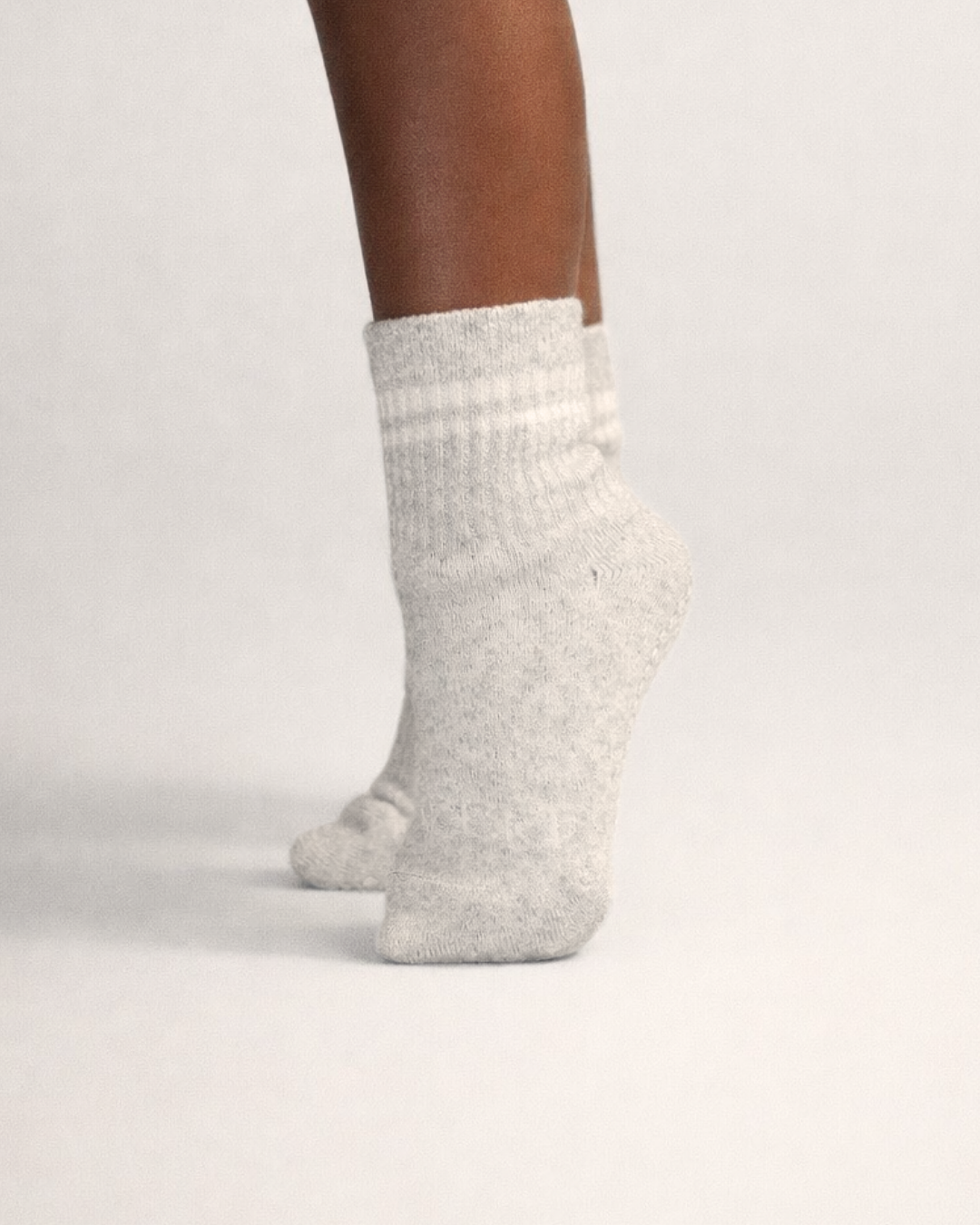 Toddler with a dark skin tone wearing a pair of heather grey toddler crew socks on a grey background.