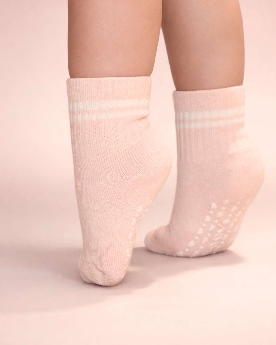 Baby with a light skin tone wearing a pair of blush baby crew socks with white heart and dot patterned silicone grips on a blush background