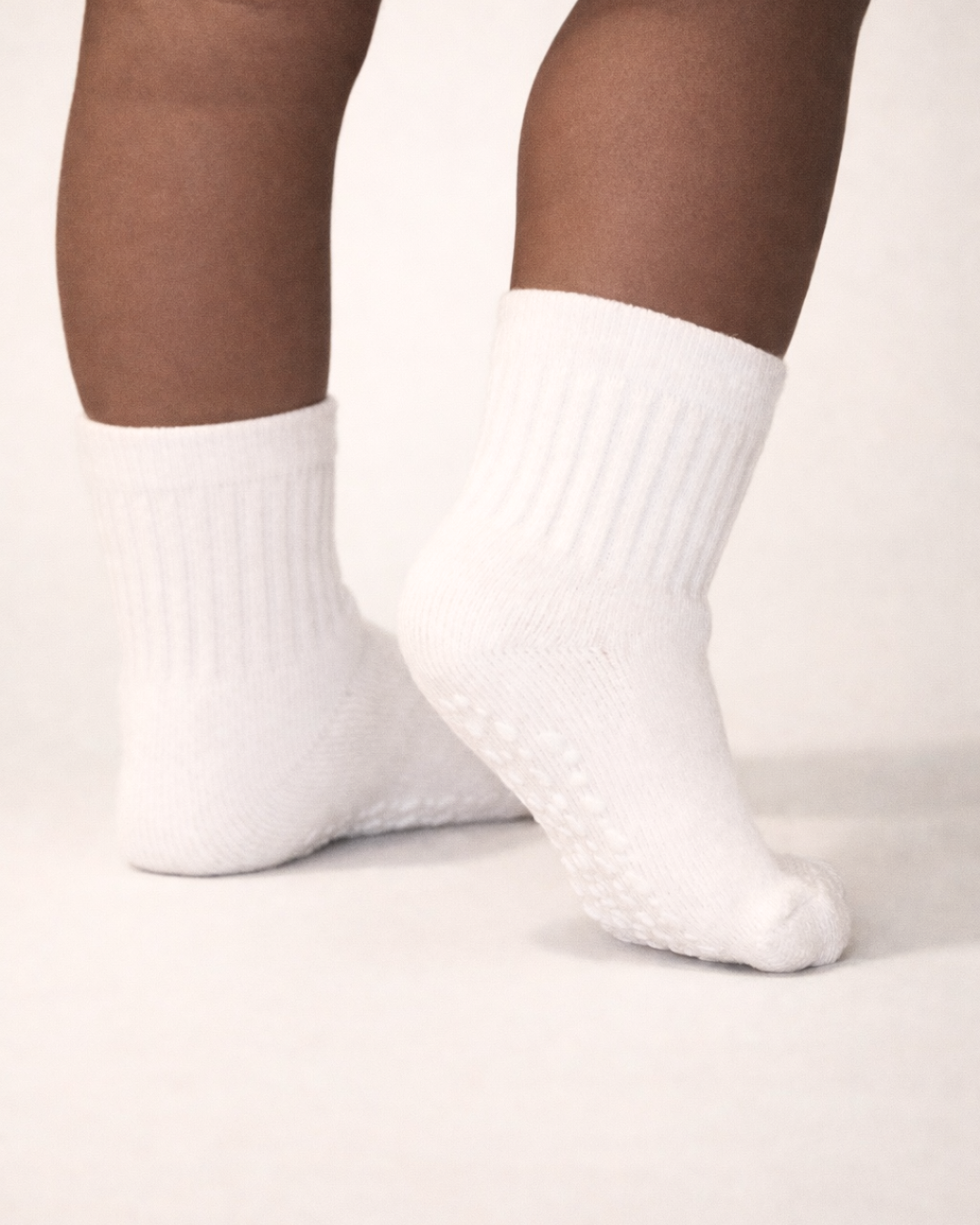 Baby with a dark skin tone wearing a pair of white baby crew socks with heart and dot patterned white silicone grips on an off-white background