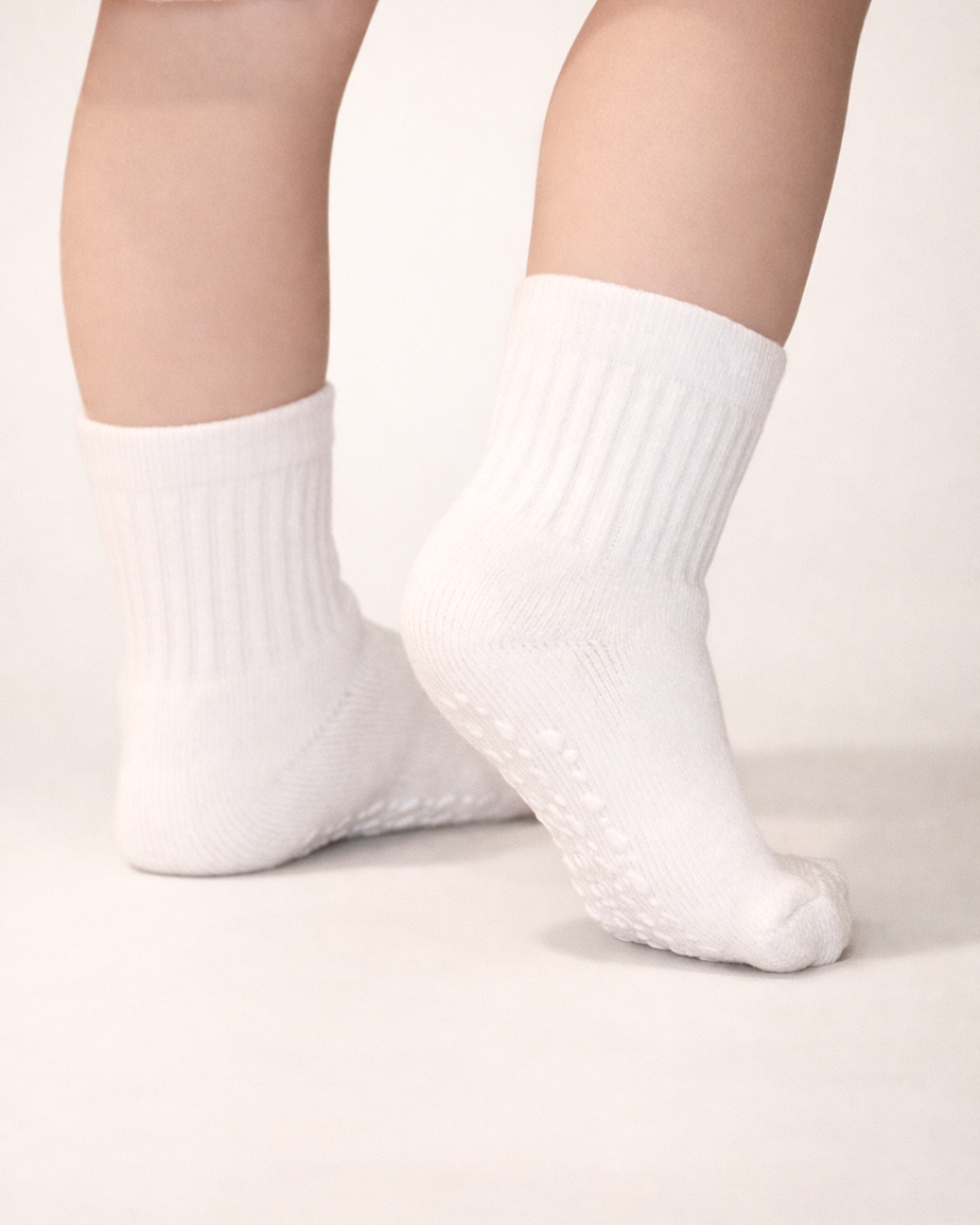 Baby with a light skin tone wearing a pair of white baby crew socks with heart and dot patterned silicone grips on an off-white background