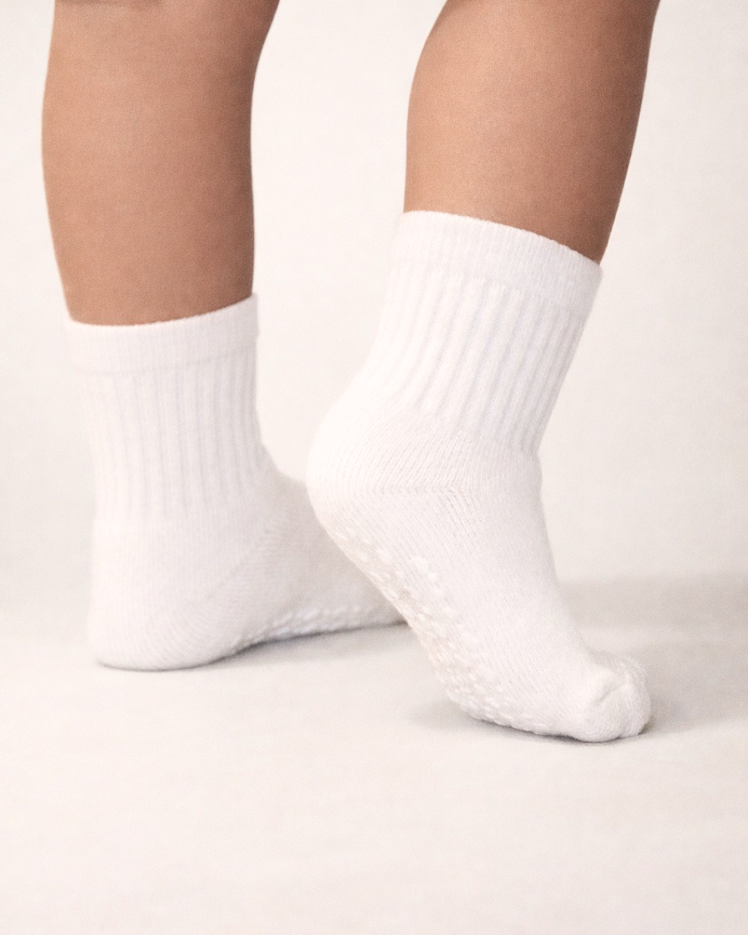 Baby with a medium skin tone wearing a pair of white baby crew socks with white heart and dot patterned silicone grips on an off-white background