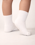 Baby with a medium skin tone wearing a pair of white baby crew socks with white heart and dot patterned silicone grips on an off-white background