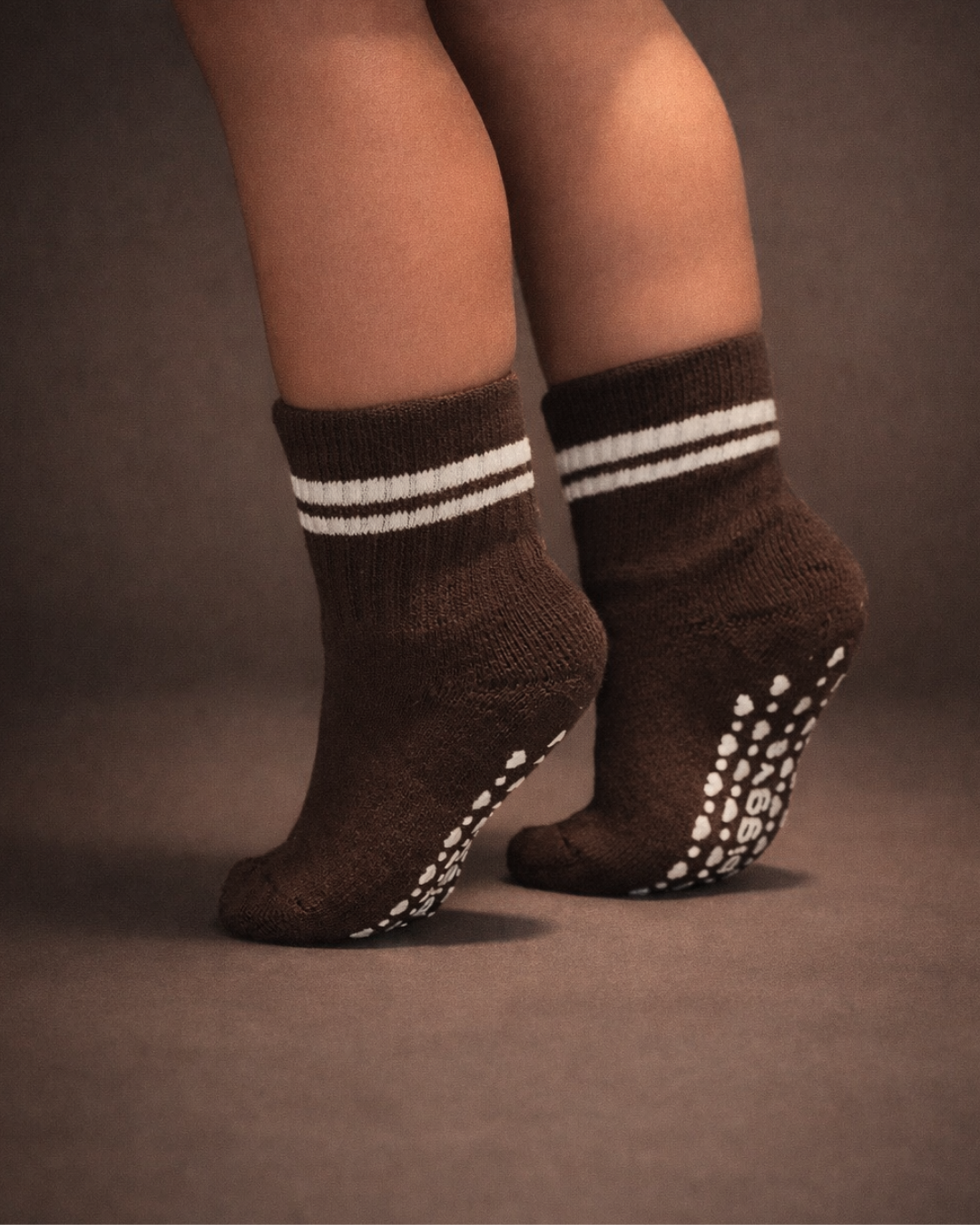 Baby with a medium skin tone wearing a pair of dark brown baby crew socks with  white silicone grips on an espresso brown background