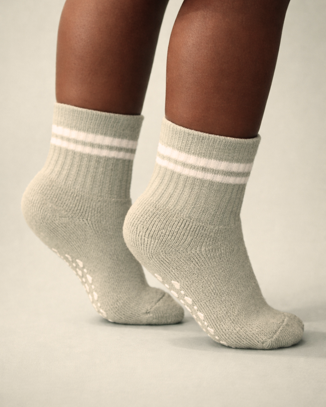 Baby with a dark skin tone wearing a pair of sage baby crew socks with white heart and dot patterned silicone grips on a sage background
