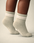 Baby with a dark skin tone wearing a pair of sage baby crew socks with white heart and dot patterned silicone grips on a sage background
