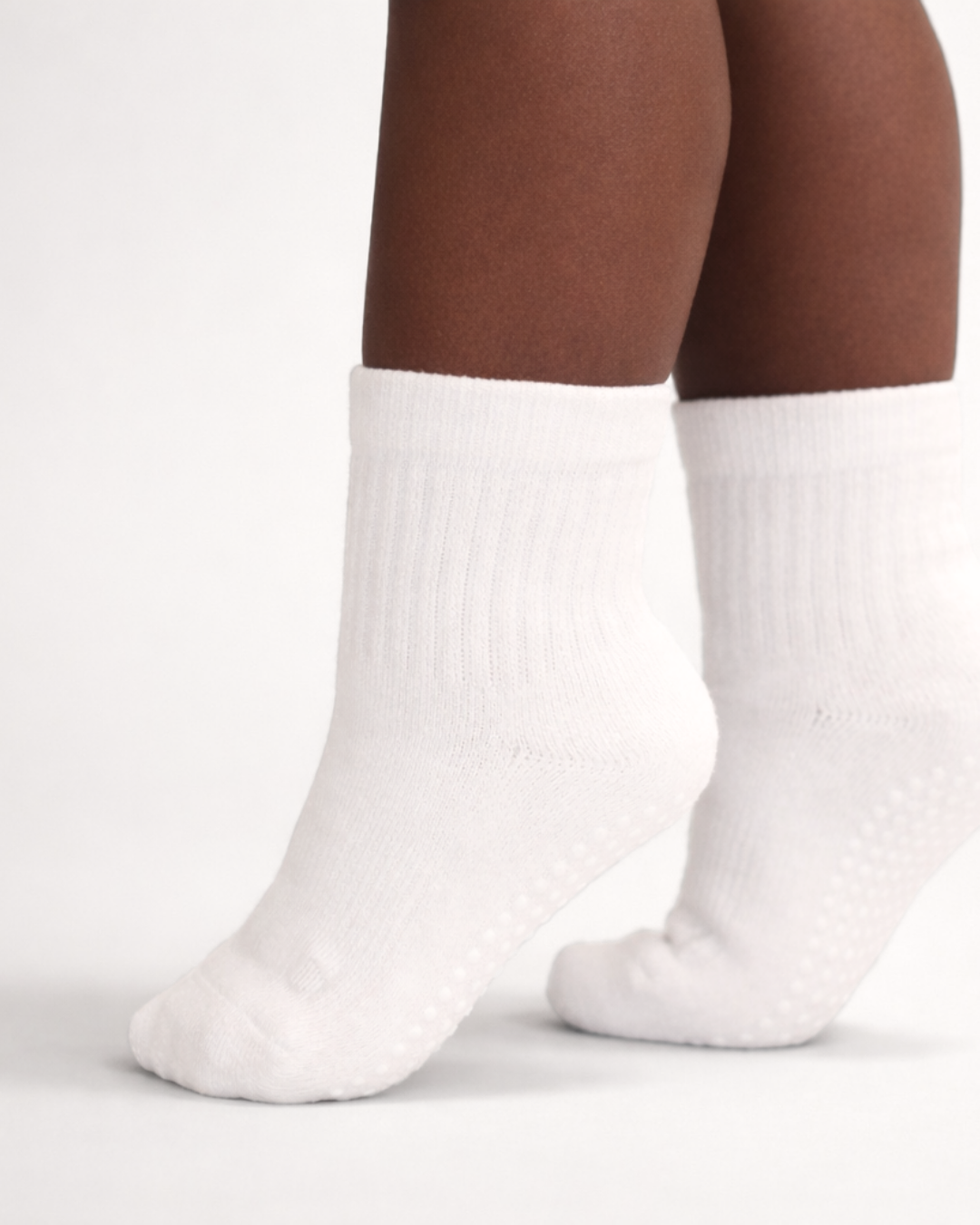 Toddler with a dark skin tone wearing a pair of white toddler crew socks on an off-white background.