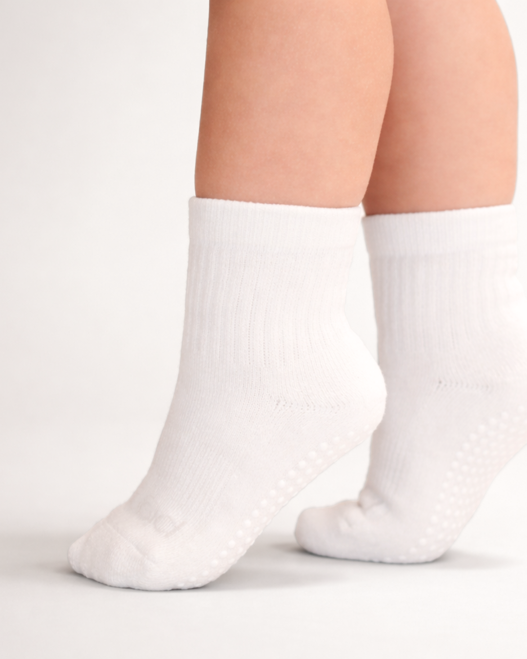 Toddler with a light skin tone wearing a pair of white toddler crew socks on an off-white background.