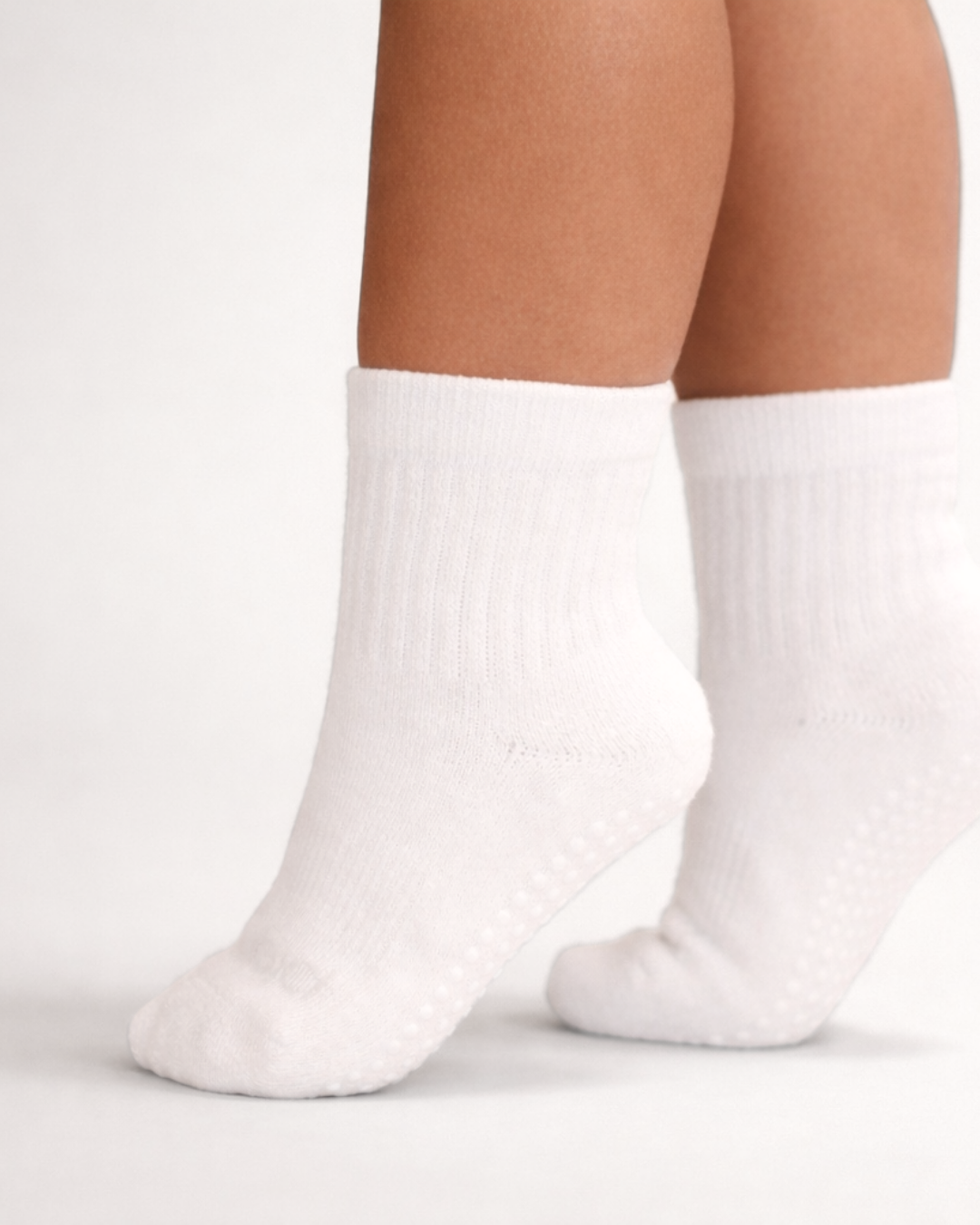 Toddler with a medium skin tone wearing a pair of white toddler crew socks on an off-white background.