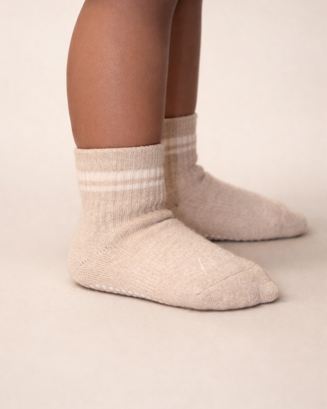 Toddler with a medium skin tone wearing a pair of heather beige toddler crew socks on a beige background.