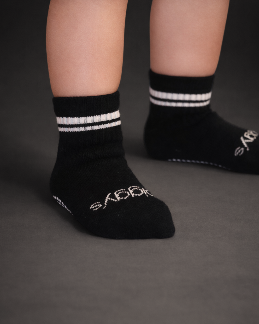 Toddler with a light skin tone wearing a pair of black toddler crew socks on a dark grey background.