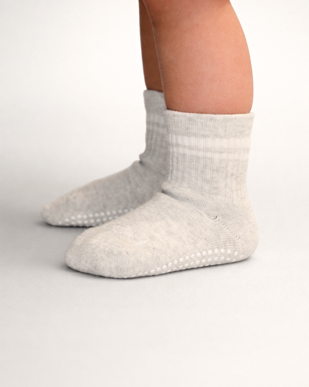 Toddler with a light skin tone wearing a pair of heather grey toddler crew socks on a grey background.