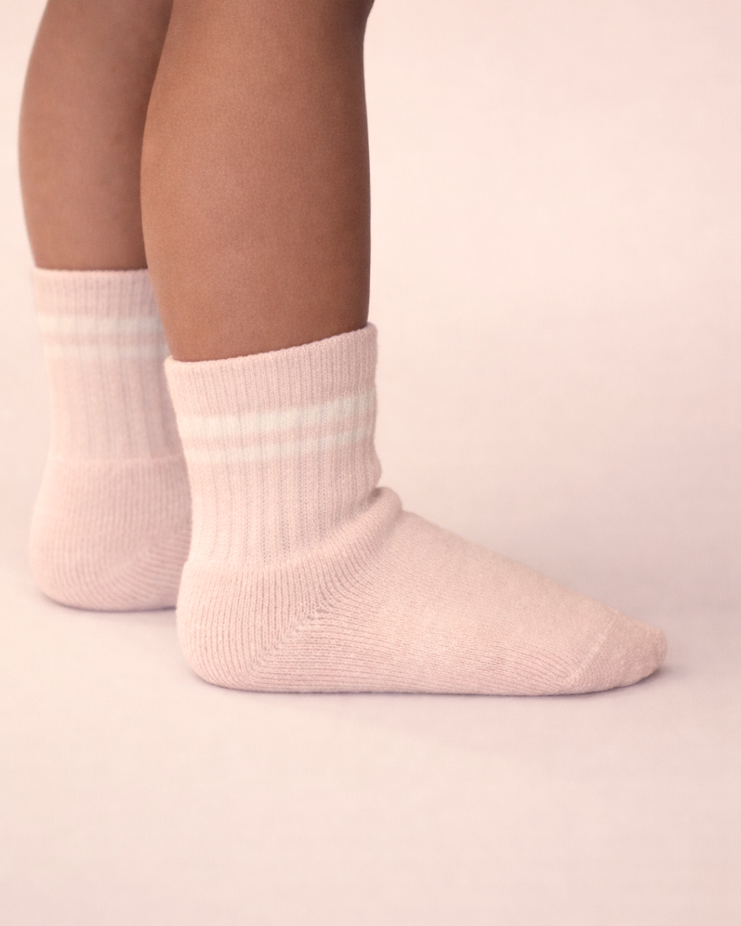 Baby with a medium skin tone wearing a pair of blush baby crew socks with white silicone grips on a blush background