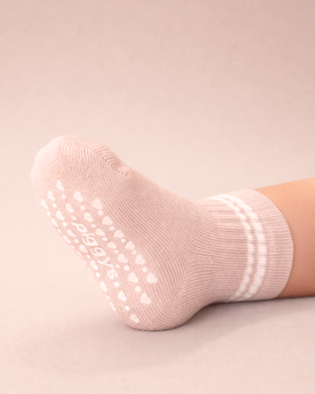 Baby with a light skin tone wearing a blush baby crew sock with white heart and dot silicone grips on a blush background