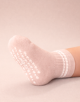 Baby with a light skin tone wearing a blush baby crew sock with white heart and dot silicone grips on a blush background