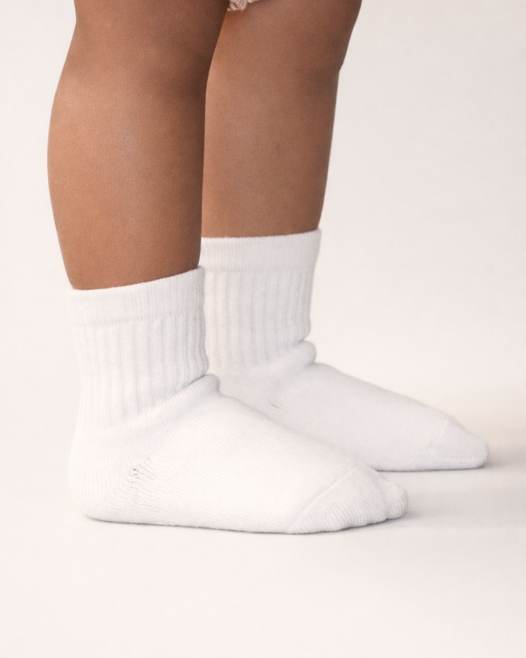 Baby with a medium skin tone wearing a pair of white baby crew socks with  silicone grips on an off-white background