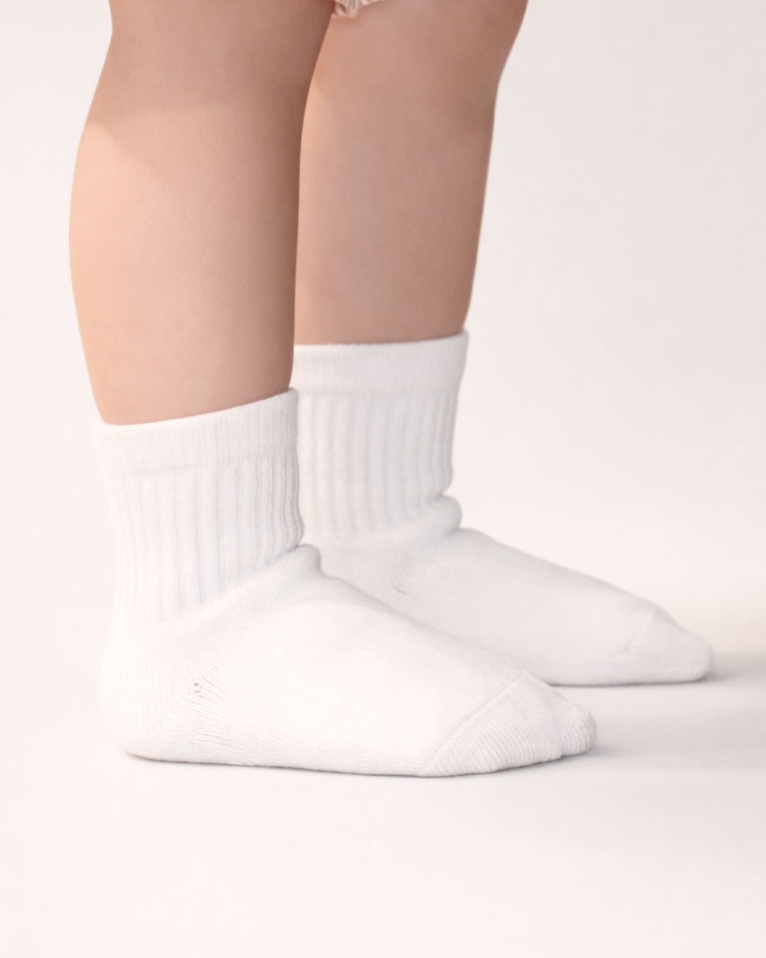 Baby with a light skin tone wearing a pair of white baby crew socks with  silicone grips on an off-white background