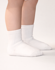 Baby with a light skin tone wearing a pair of white baby crew socks with  silicone grips on an off-white background