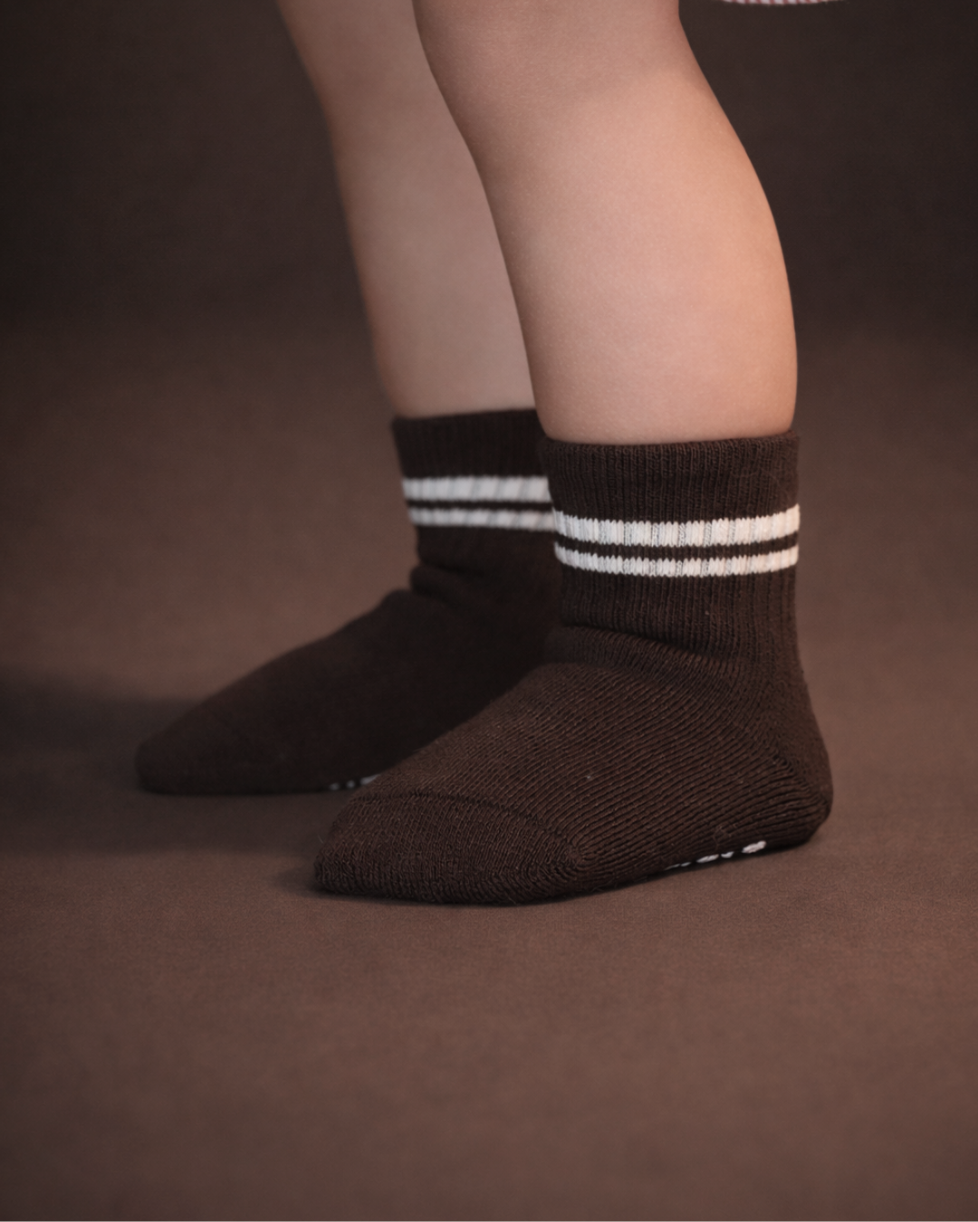 Baby with a light skin tone wearing a pair of dark brown baby crew socks with  white silicone grips on an espresso brown background