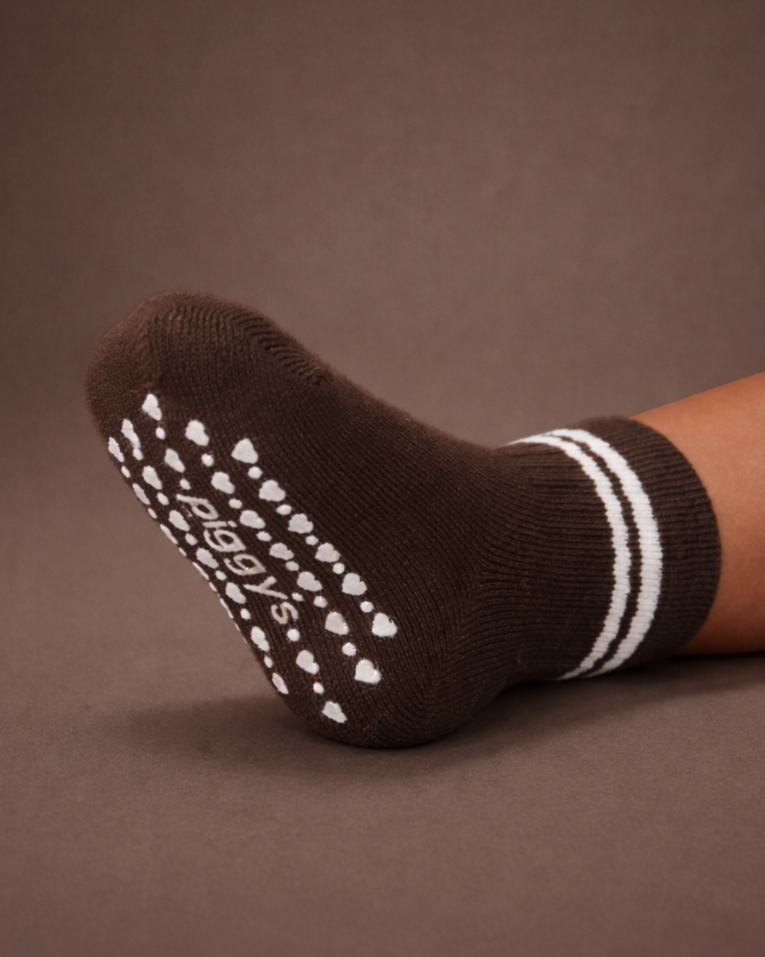 Baby with a medium skin tone wearing an espresso brown baby crew sock with  white heart and dot silicone grips on an espresso brown background